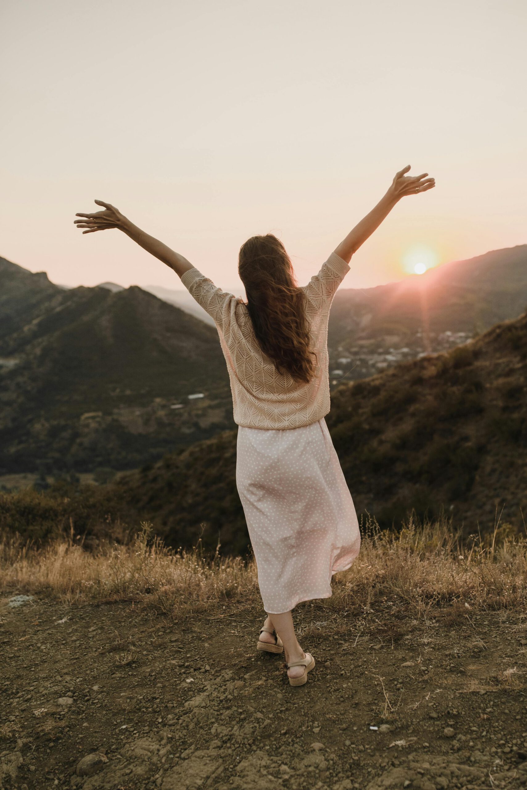 A woman with arms raised stands on a mountain trail, embracing the sunset view in an outdoor landscape.